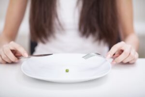 Hands of a woman holding a knife and fork poised above a plate with a single pea, symbolizing restrictive eating habits, featured in a guide exploring types of eating disorders.