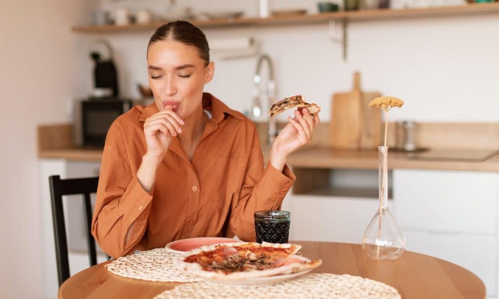 Satisfied woman licking her fingers after enjoying a slice of pizza, representing the enjoyment of food in discussions about early intervention and awareness of eating disorders.
