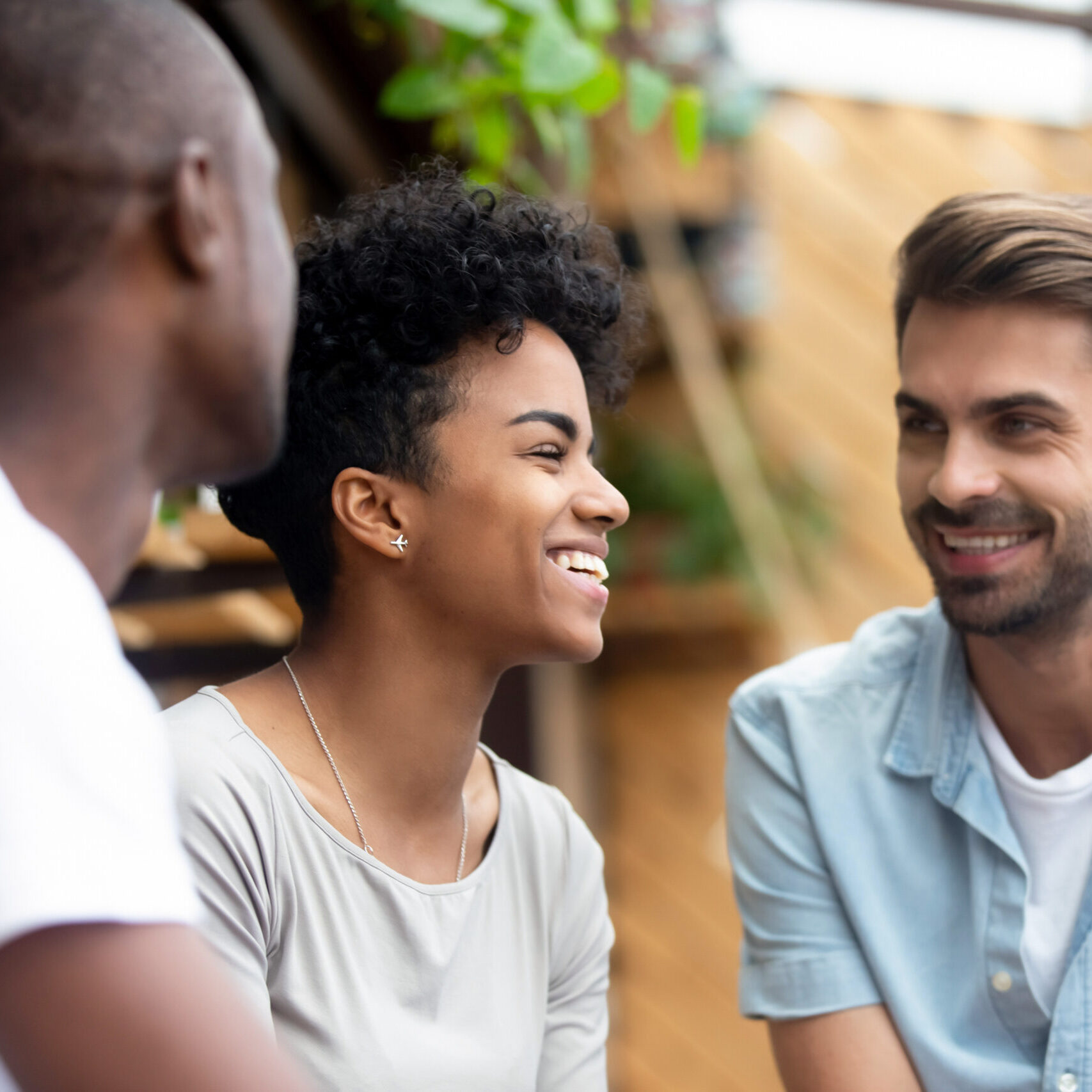 Three millennial multi-ethnic friends spending weekend together sitting in cafe or restaurant cozy summer terrace talking enjoying free time with each other, focus on laughing attractive african girl