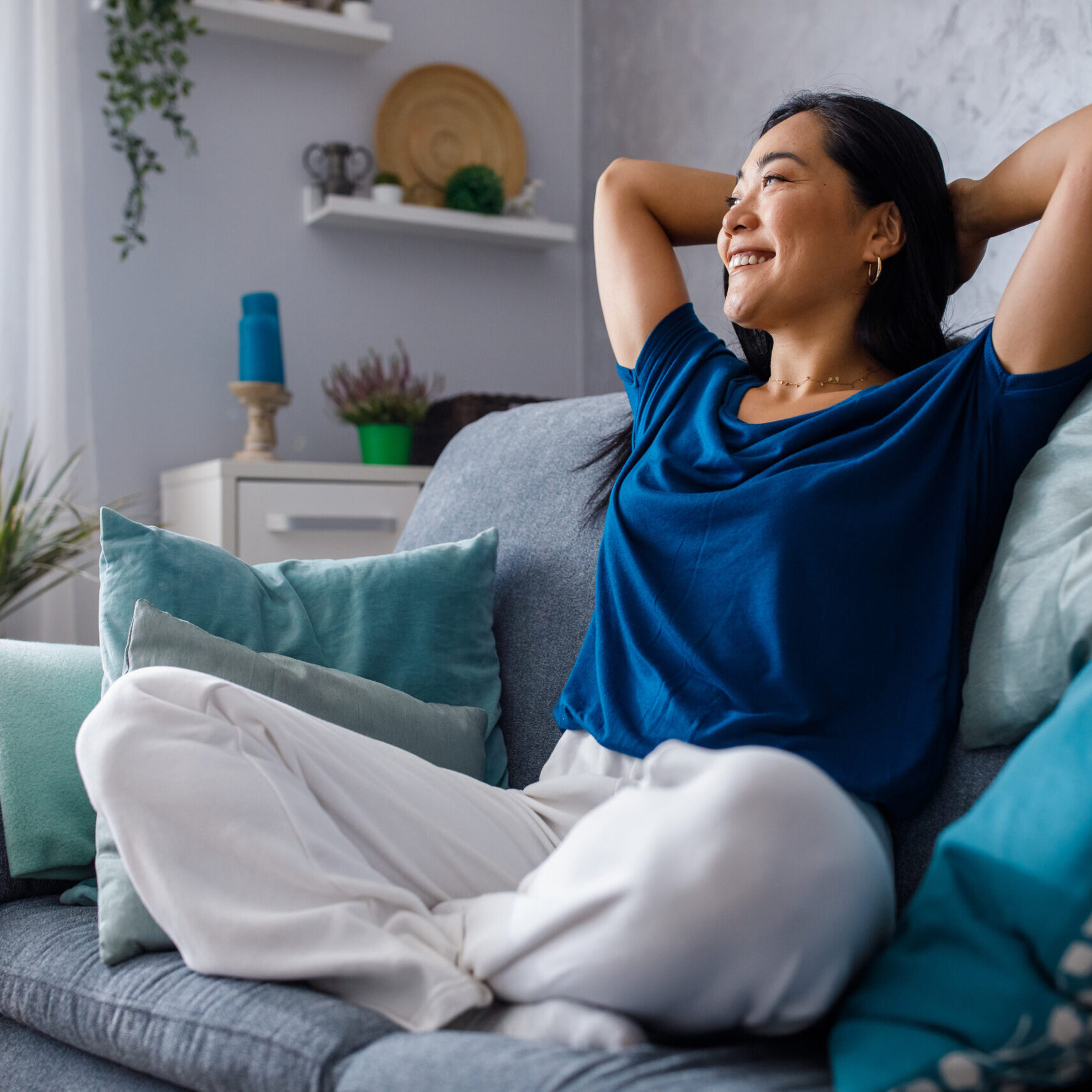 Full length shot of beautiful, happy young woman sitting on the cozy sofa with hands behind head, looking away, smiling and daydreaming.